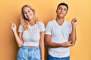 Young interracial couple wearing casual white tshirt with a big smile on face, pointing with hand and finger to the side looking at the camera.