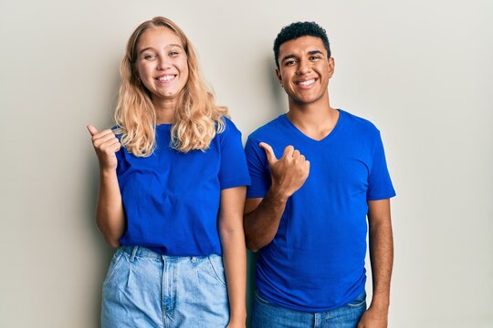 Young interracial couple wearing casual clothes pointing to the back behind with hand and thumbs up, smiling confident