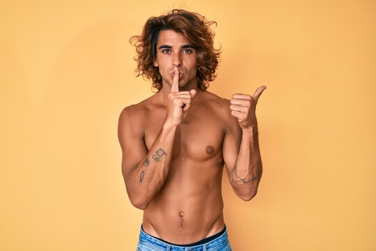 Young hispanic man standing shirtless asking to be quiet with finger on lips pointing with hand to the side. silence and secret concept.