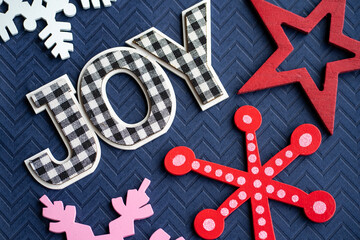 Christmas wood snowflake background, with diagonal JOY letters in black and white plaid, on blue background. Colors of pink, red and white