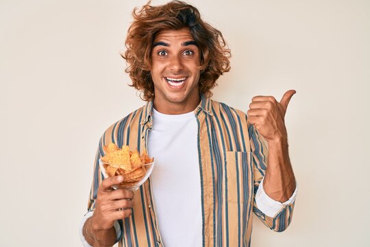Young hispanic man holding nachos potato chips pointing thumb up to the side smiling happy with open mouth