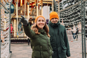 Follow me shot of a young attractive loving couple at the Christmas market in winter. Man and woman wearing orange hats and green jackets walking among garland lights outdoors. Holidays concept.