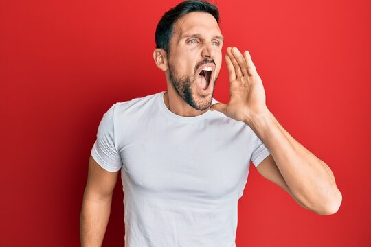 Young handsome man wearing casual white tshirt shouting and screaming loud to side with hand on mouth. communication concept.