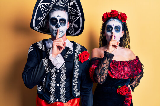 Young couple wearing mexican day of the dead costume over yellow asking to be quiet with finger on lips. silence and secret concept.