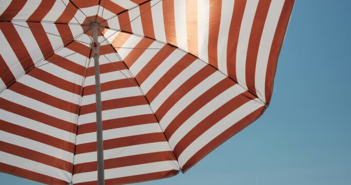 Sky shot POV of a red stripe umbrella on a solid blue sky creating shadow on a beach. Sunny day on vacation. Balanced commercial composition. Relaxation light chill holiday mood vibe colorful 4k video