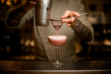 male bartender filters whipped cocktail through sieve into wine glass on bar counter