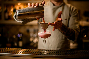 male bartender holds sieve over wine glass and gently pours cocktail from shaker.
