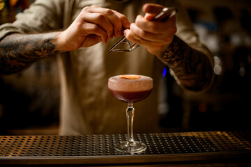 male bartender accurate grates cinnamon over glass of cocktail with foam