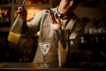 view on barman pours drink from metal jigger into shaker on bar counter