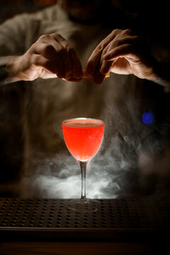 Bartender Holding Lemon Peel And Sprinkles On Glass With Cocktail At Smoky Bar.