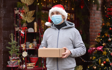 Safe delivery of gifts and packages for the holidays in quarantine during the coronavirus pandemic. A courier, a volunteer in a Santa hat, a medical mask and gloves, holds a box.Copy space for text