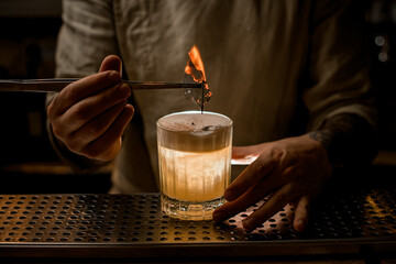 Fototapeta na wymiar bartender holds tweezers with burning leaf to decorate glass of cocktail