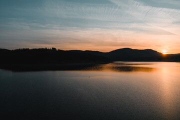 Fototapeta na wymiar A beautiful sunset at a mountain lake with mountain silhouettes and warm orange light. Grane Dam, Granetalsperre, Harz Mountains
