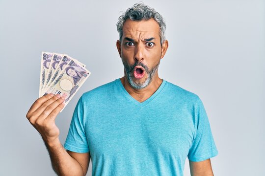 Middle age grey-haired man holding japanese yen banknotes scared and amazed with open mouth for surprise, disbelief face