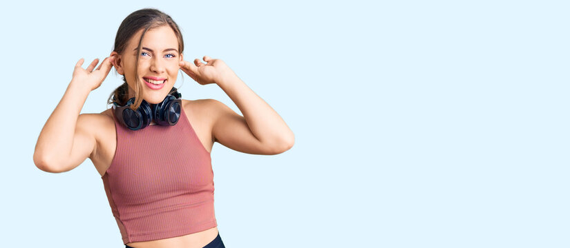 Beautiful caucasian young woman wearing gym clothes and using headphones smiling pulling ears with fingers, funny gesture. audition problem