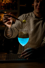 male bartender at dark bar holds golden piece of ice over steaming wine glass