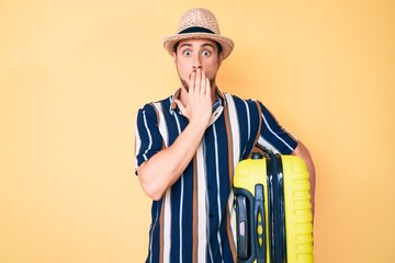 Obraz premium Young handsome man wearing summer hat holding cabin bag covering mouth with hand, shocked and afraid for mistake. surprised expression