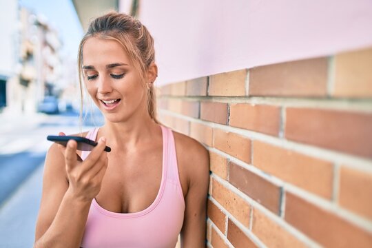 Young blonde sporty girl talking on the smartphone leaning on the wall at street of city.