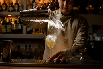 barman pours steaming drink from steel shaker cup into wine glass with ice cubes.