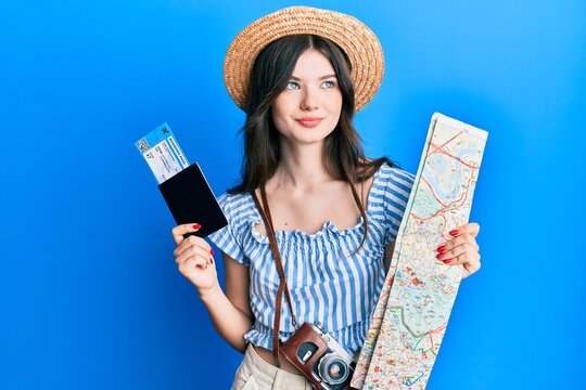 Young beautiful caucasian girl holding passport and city map smiling looking to the side and staring away thinking.