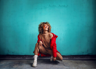 Young fashionable smiling woman with curly hair, in red jacket crouching in front of the wall and looking away.