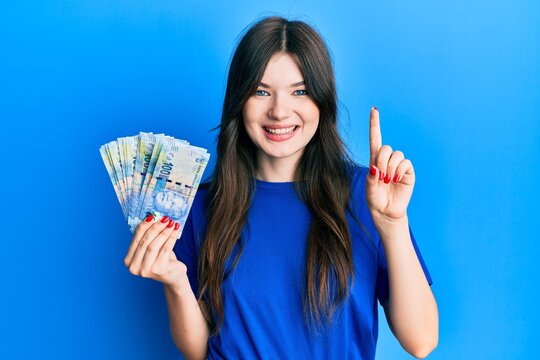 Young beautiful caucasian girl holding south african 100 rand banknotes smiling with an idea or question pointing finger with happy face, number one