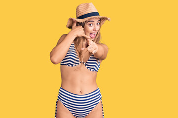 Young beautiful blonde woman wearing bikini and hat smiling doing talking on the telephone gesture...