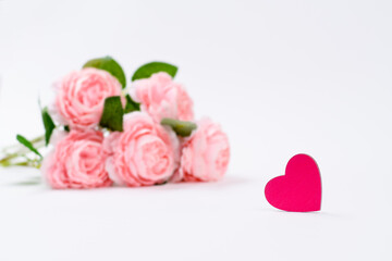 Pink heart and a bouquet of pink flowers on a white background. Focusing on the heart, the roses in the background are blurred.