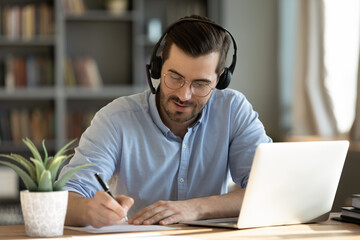 Focused 35s man sit at desk wear headphones watch webinar use laptop gain new knowledge writing...