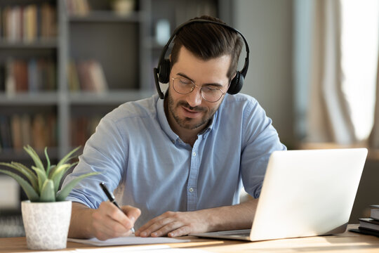 Focused 35s man sit at desk wear headphones watch webinar use laptop gain new knowledge writing notes. Video conference communication, negotiation remotely, study online, e-learn, self-educate concept
