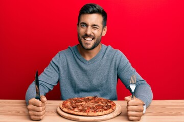 Handsome hispanic man eating tasty pepperoni pizza winking looking at the camera with sexy expression, cheerful and happy face.