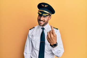 Handsome hispanic man wearing airplane pilot uniform beckoning come here gesture with hand inviting welcoming happy and smiling