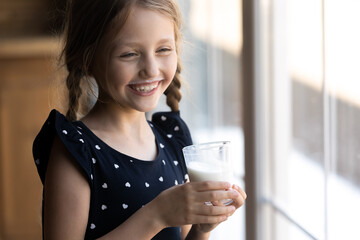 I have got mustache. Naughty little girl standing by window drinking tasty dairy products fresh milk or natural yoghurt from glass, laughing while understanding she has funny white whiskers under nose