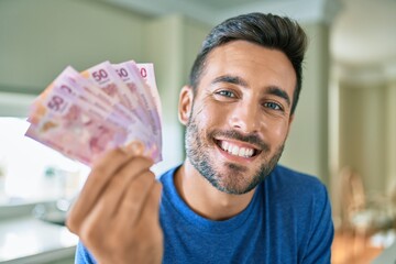 Young handsome man smiling happy holding mexican pesos banknotes at home - obrazy, fototapety, plakaty