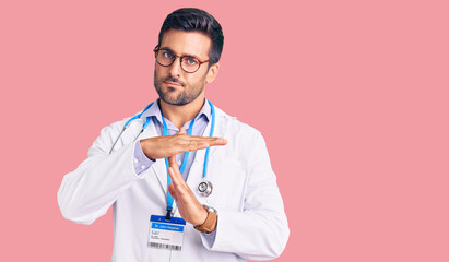 Young hispanic man wearing doctor uniform and stethoscope doing time out gesture with hands,...