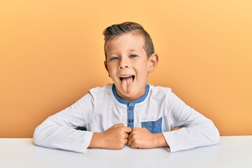 Adorable caucasian kid wearing casual clothes sitting on the table sticking tongue out happy with...