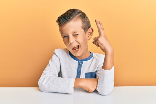 Adorable caucasian kid wearing casual clothes sitting on the table smiling with happy face winking at the camera doing victory sign. number two.