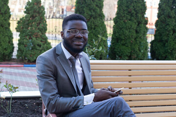 Obraz premium Young African businessman smiling on city bench with phone, tablet