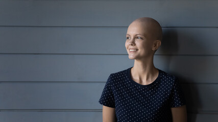 Never give up. Young smiling woman cancer patient having bald head after chemotherapy standing near grey wooden wall looking at distance expressing optimism motivation courage hope belief, copy space
