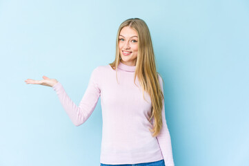 Young blonde woman isolated on blue background showing a copy space on a palm and holding another hand on waist.