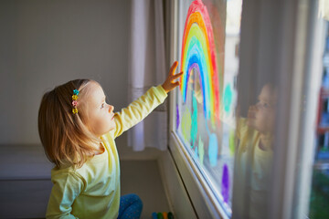 Adorable toddler girl drawing rainbow on window glass