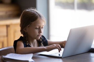 Distant learning. Concentrated little girl sitting at desk at home typing on keyboard of laptop...