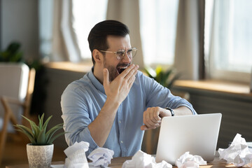 Exhausted 35s businessman sit at desk cover mouth with hand while yawning looks at wristwatch feels...
