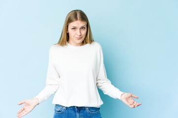 Young blonde woman isolated on blue background shrugs shoulders and open eyes confused.