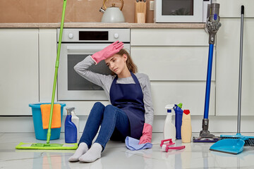 A tired, exhausted young woman after a long cleaning of the kitchen is sitting on the floor. Nearby equipment for cleaning the house. Cleaning the house.
