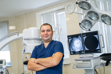 Obraz premium Portrait of an handsome doctor standing in hospital room. Medic smiles to the camera with crossed hands.