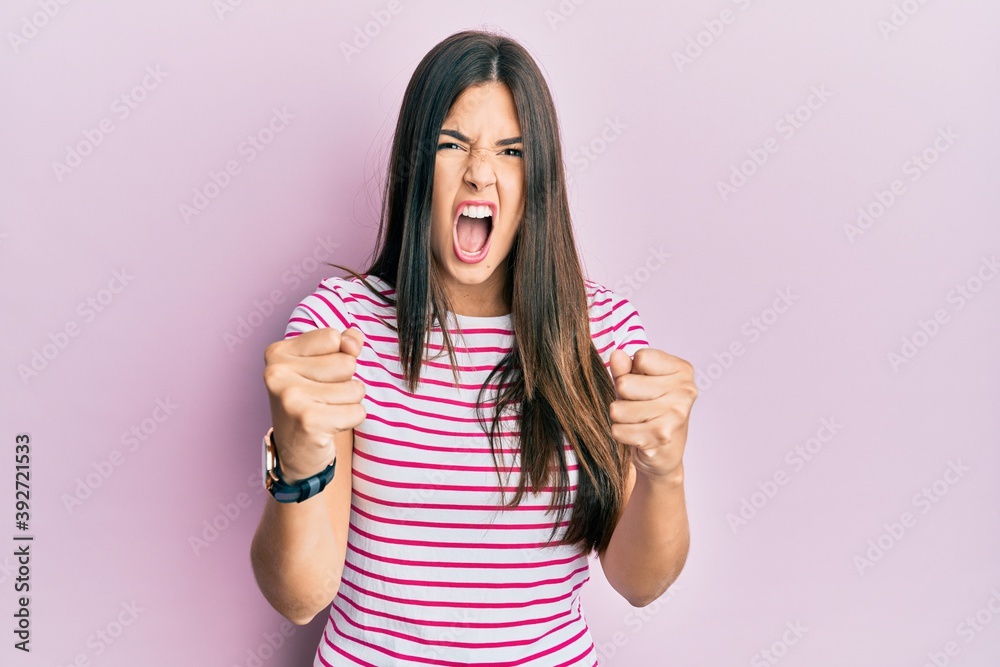 Sticker Young brunette woman wearing casual clothes over pink background angry and mad raising fists frustrated and furious while shouting with anger. rage and aggressive concept.