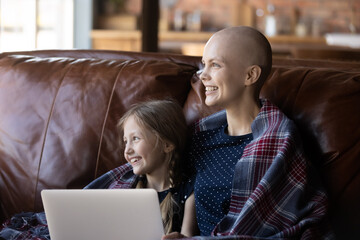 Cozy evening at home. Little girl and loving millennial mother cancer patient sitting on couch...