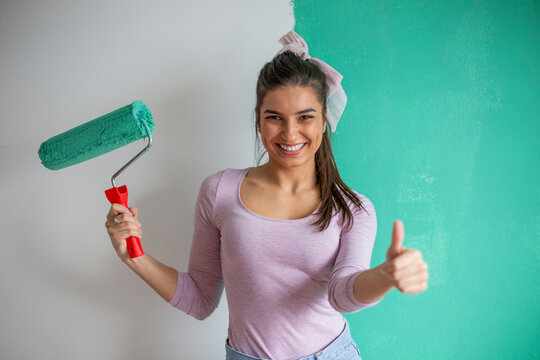 Portrait of smiling young woman painting an interior wall with a paint roller and showing thumb up.