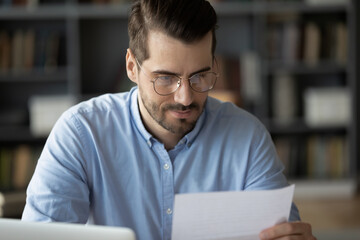 Serious focused man sitting at workplace desk holding paper sheet reading news in correspondence...